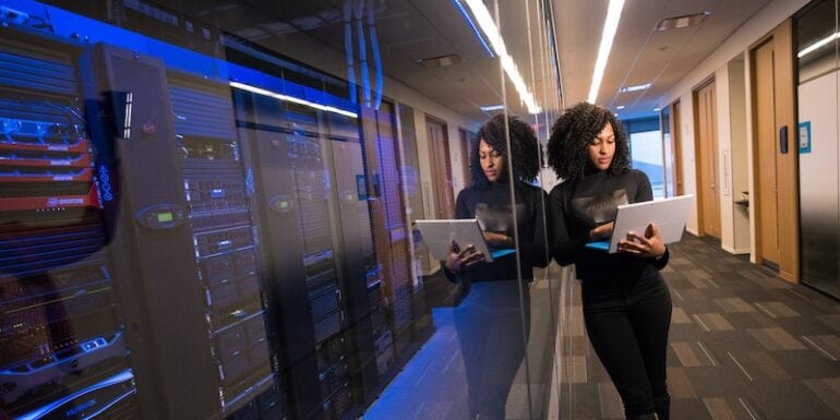 A woman on a laptop leaning against a glass wall with servers behind it.