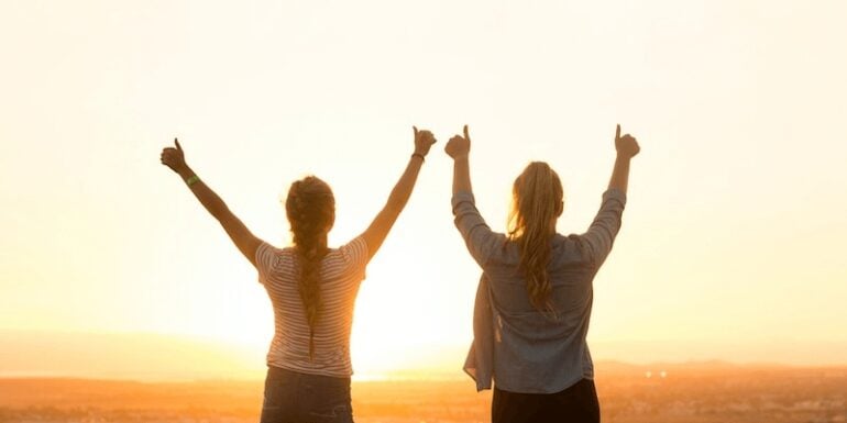 Women standing with their thumbs up at sunset.